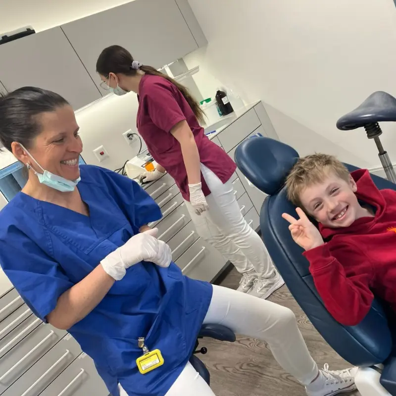 Kind sitzt entspannt im Zahnarztstuhl und lächelt nach der Behandlung beim Zahnarzt, macht ein peace Zeichen